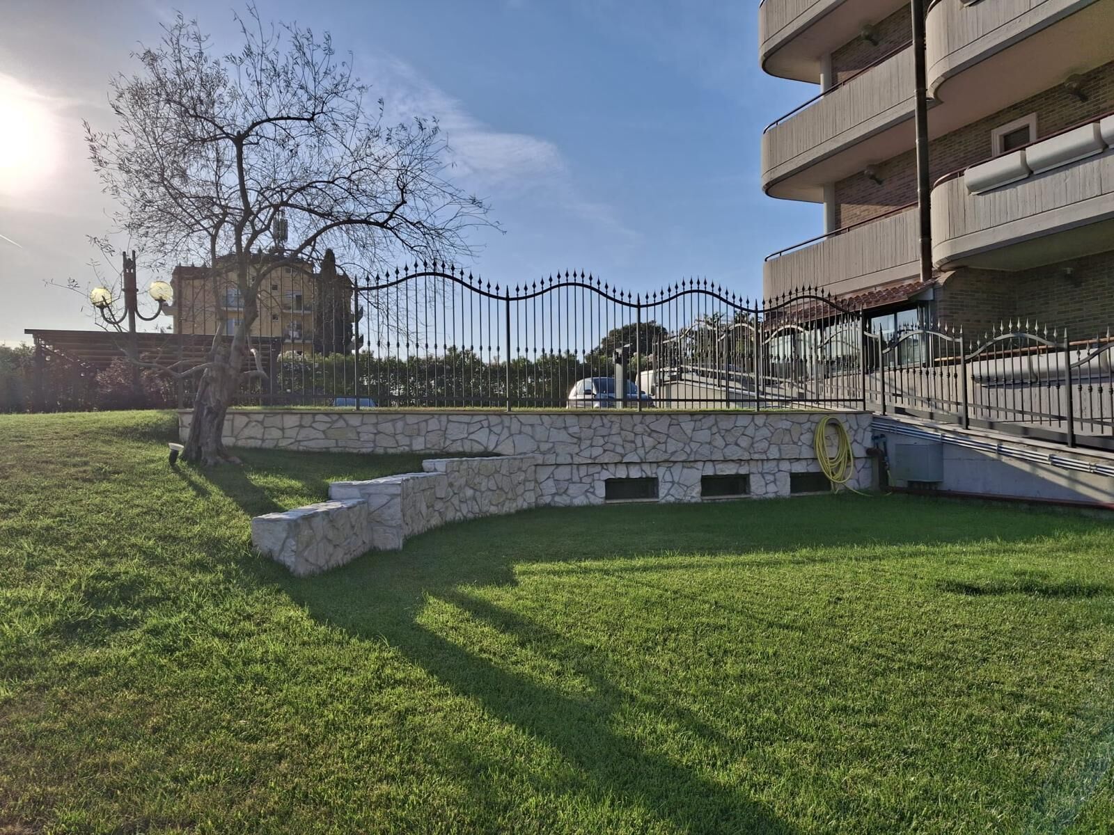 Un patio con césped, un muro de piedra, una cerca metálica y un edificio con balcones.