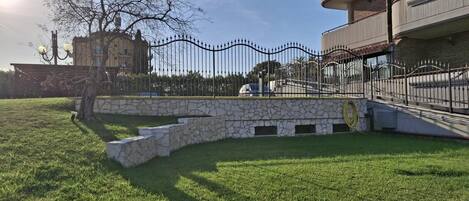 Un patio con césped, un muro de piedra, una cerca metálica y un edificio con balcones.