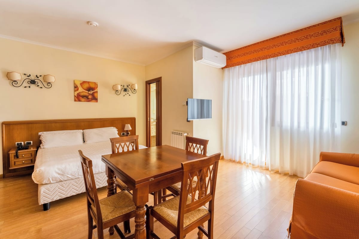 A hotel room with a wooden bed, a dining table with chairs, a TV, and a large window with curtains.