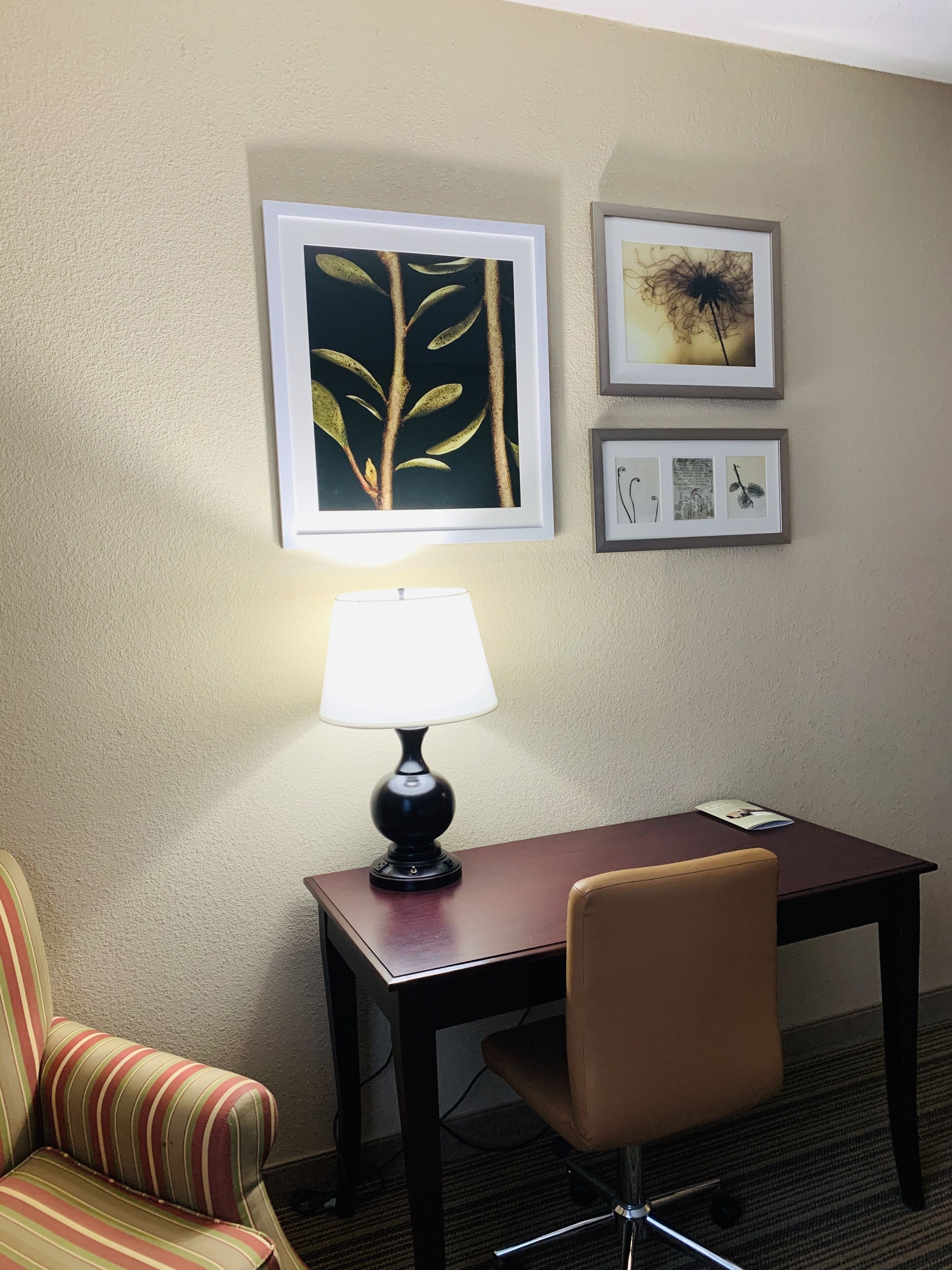 A hotel room with a desk, a chair, a lamp, and three framed artworks on the wall.