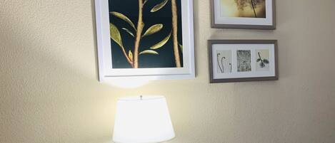 A hotel room with a desk, a chair, a lamp, and three framed artworks on the wall.