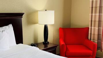 A hotel room with a bed, a red armchair, a nightstand with a lamp, and a window with curtains.