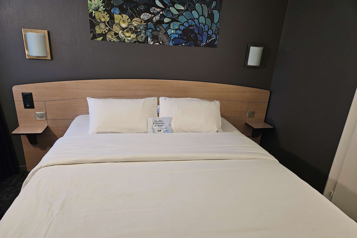 Una habitación de hotel con una cama, dos mesitas de noche, una lámpara de noche, una obra de arte en la pared y una puerta.