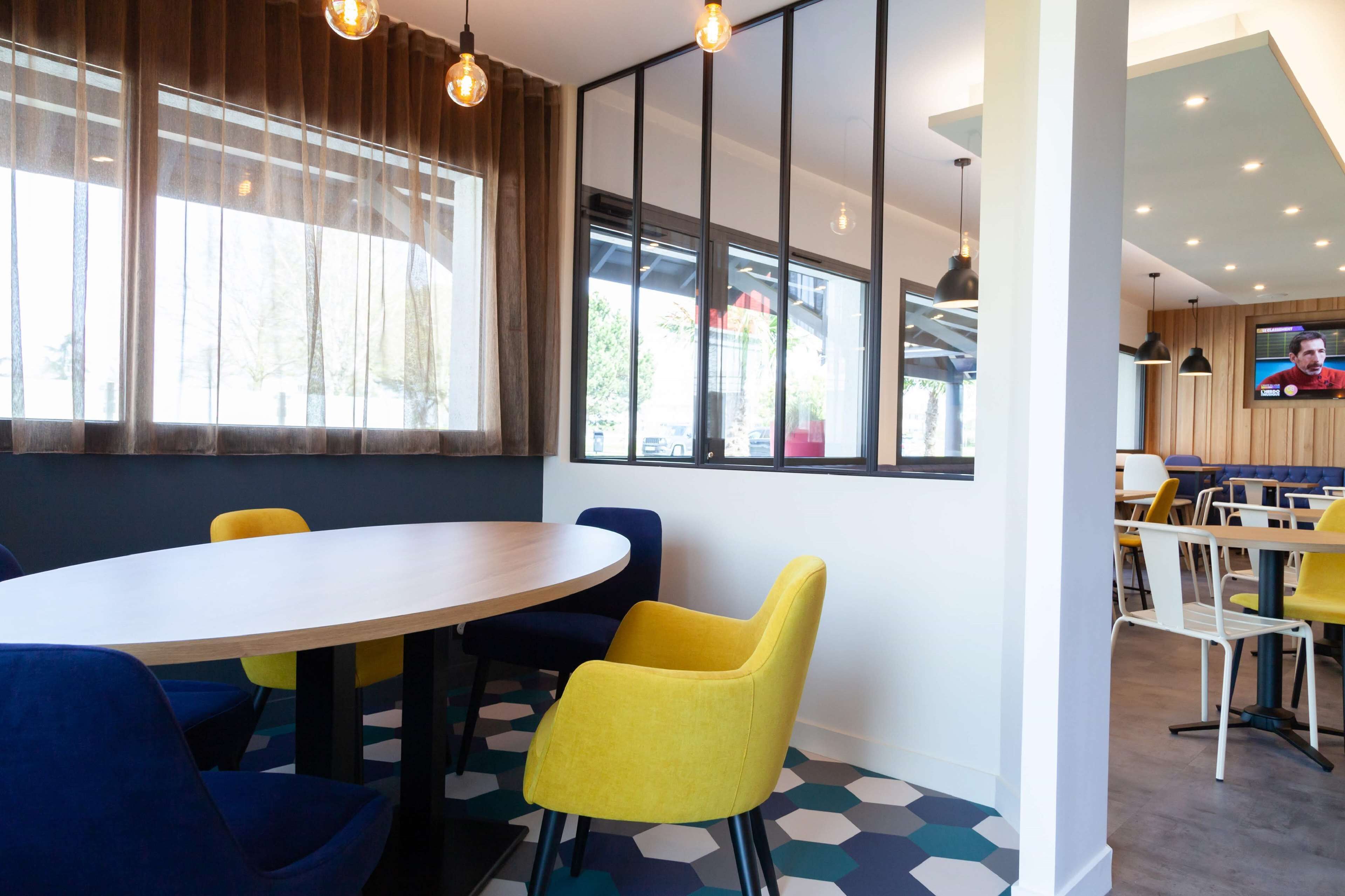 A modern dining area with round tables and colorful chairs, large windows with curtains, and a television on the wall.