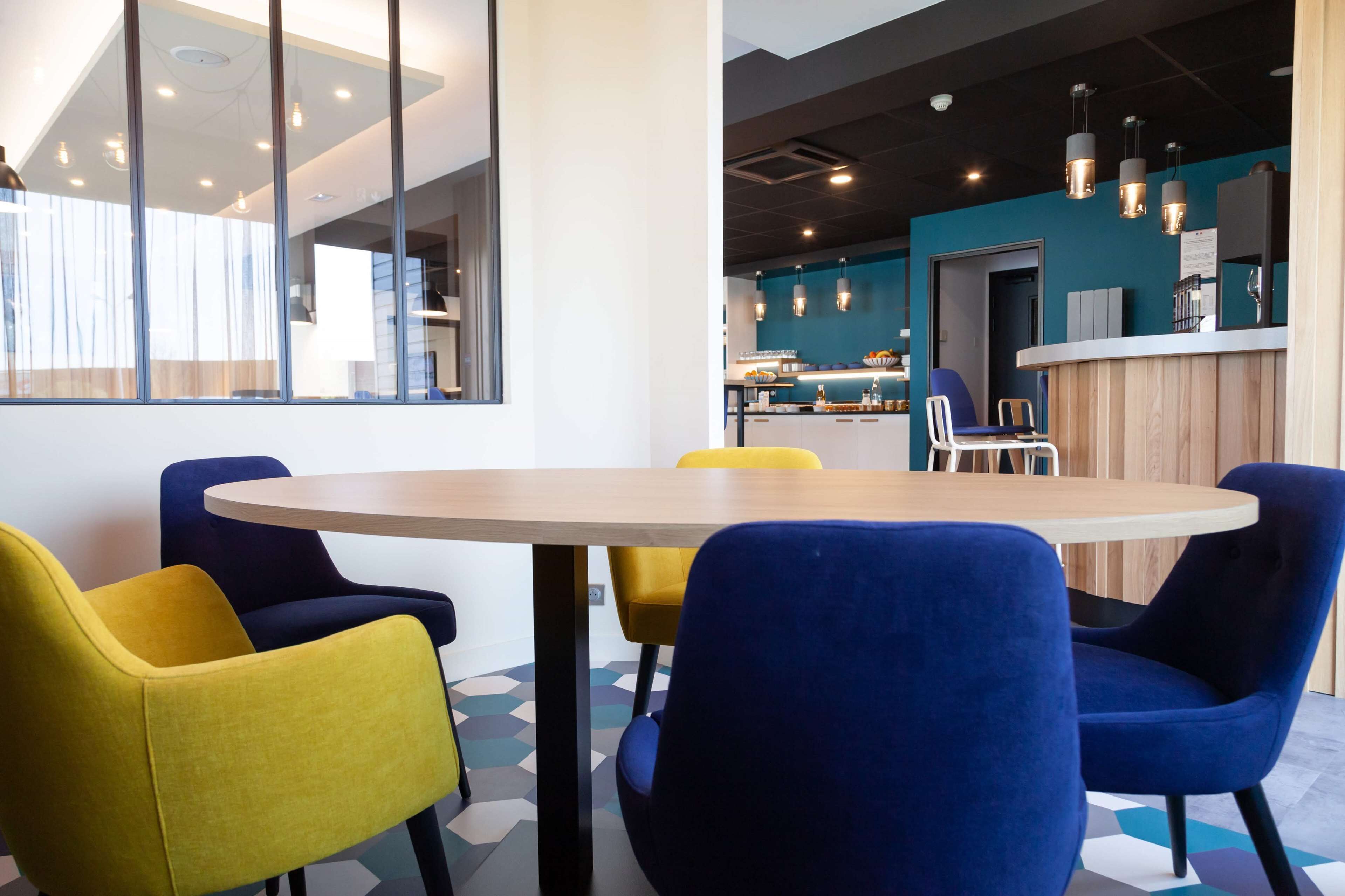 A modern meeting room with a round table and colorful chairs, a reception area with a wooden counter, and a kitchen in the background.