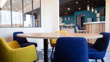 A modern meeting room with a round table and colorful chairs, a reception area with a wooden counter, and a kitchen in the background.