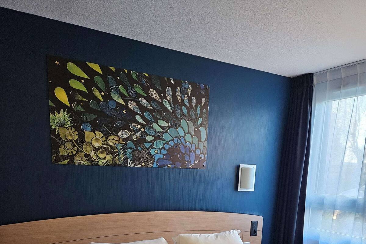 Une chambre d’hôtel avec un grand lit, une tête de lit, une lampe fixée au mur et une fenêtre avec des rideaux.