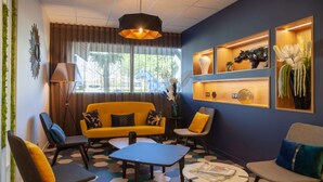 Un moderno área de descanso con un sofá amarillo, una mesa de centro azul y una variedad de sillones. Hay estanterías con objetos decorativos y un ventanal con cortinas.
