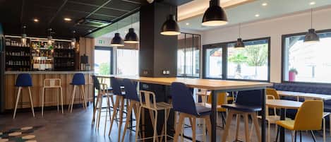 A modern bar area with wooden tables and chairs, a bar counter with stools, and pendant lighting.