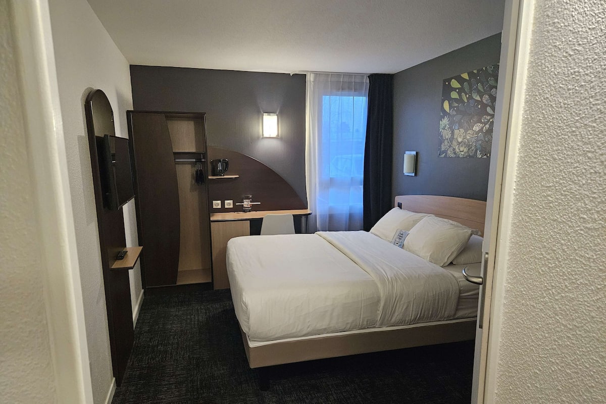 Une chambre d’hôtel équipée d’un lit, d’un bureau, d’un miroir, d’une petite fenêtre avec des rideaux et d’une lampe fixée au mur.