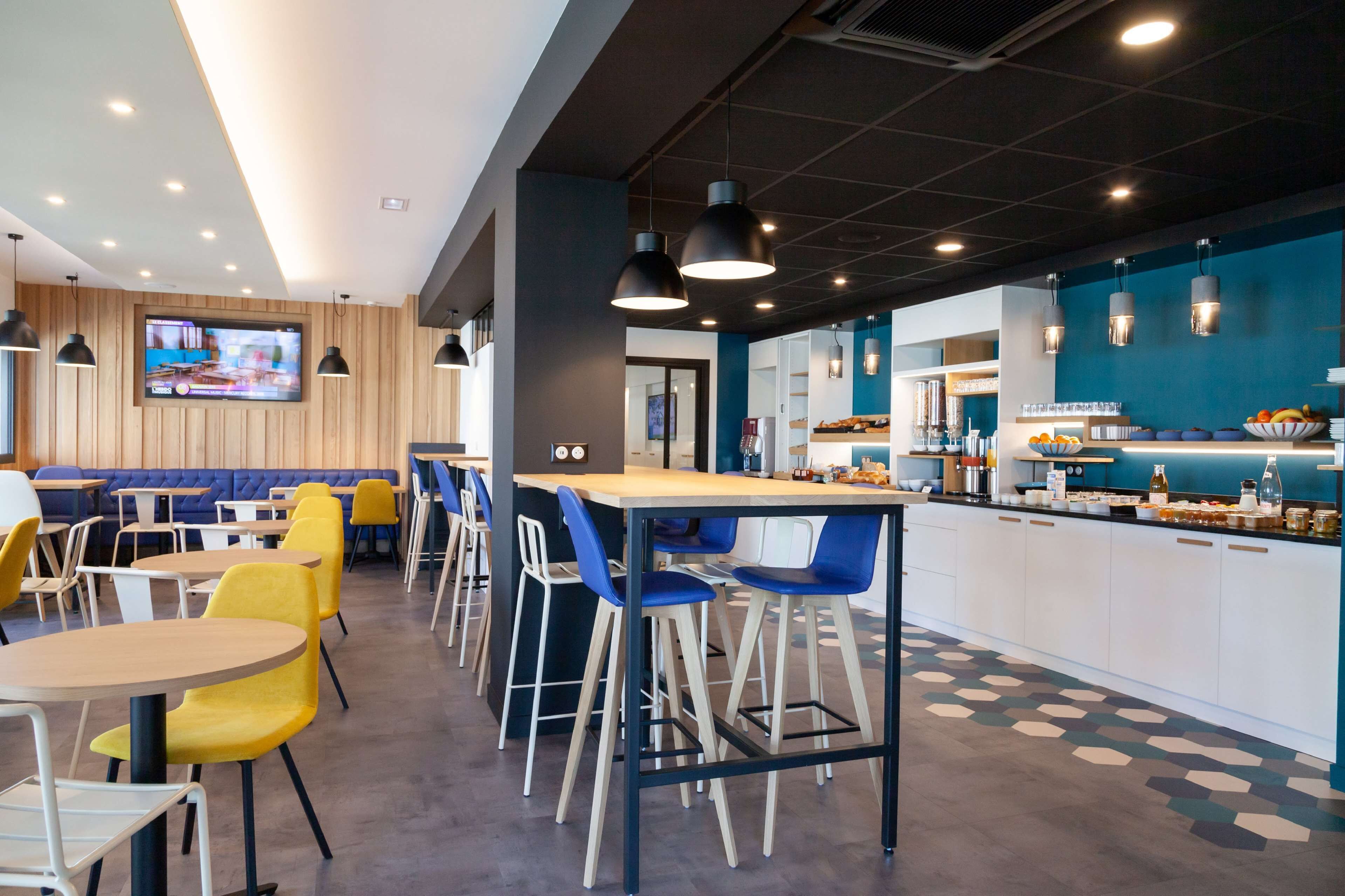 A modern cafe with a long counter, blue and yellow chairs, and a television mounted on the wall.