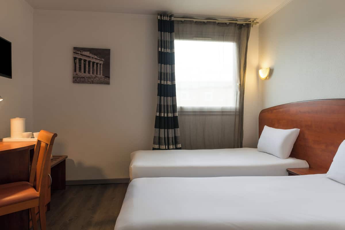 A hotel room with two beds, a desk with a chair, a window with curtains, and a wall-mounted picture.