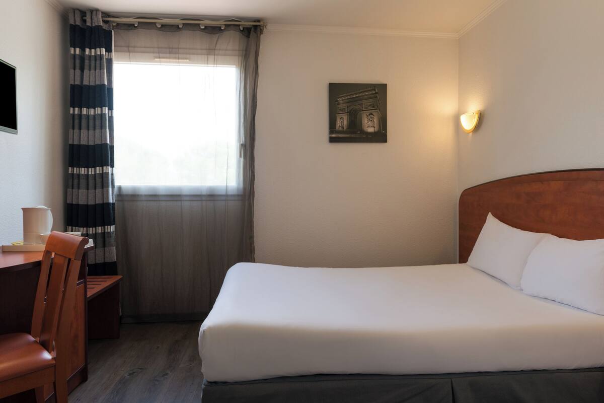 A hotel room with a bed, a desk, a chair, a window with curtains, and a wall-mounted light.