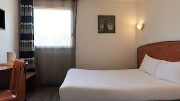 A hotel room with a bed, a desk, a chair, a window with curtains, and a wall-mounted light.