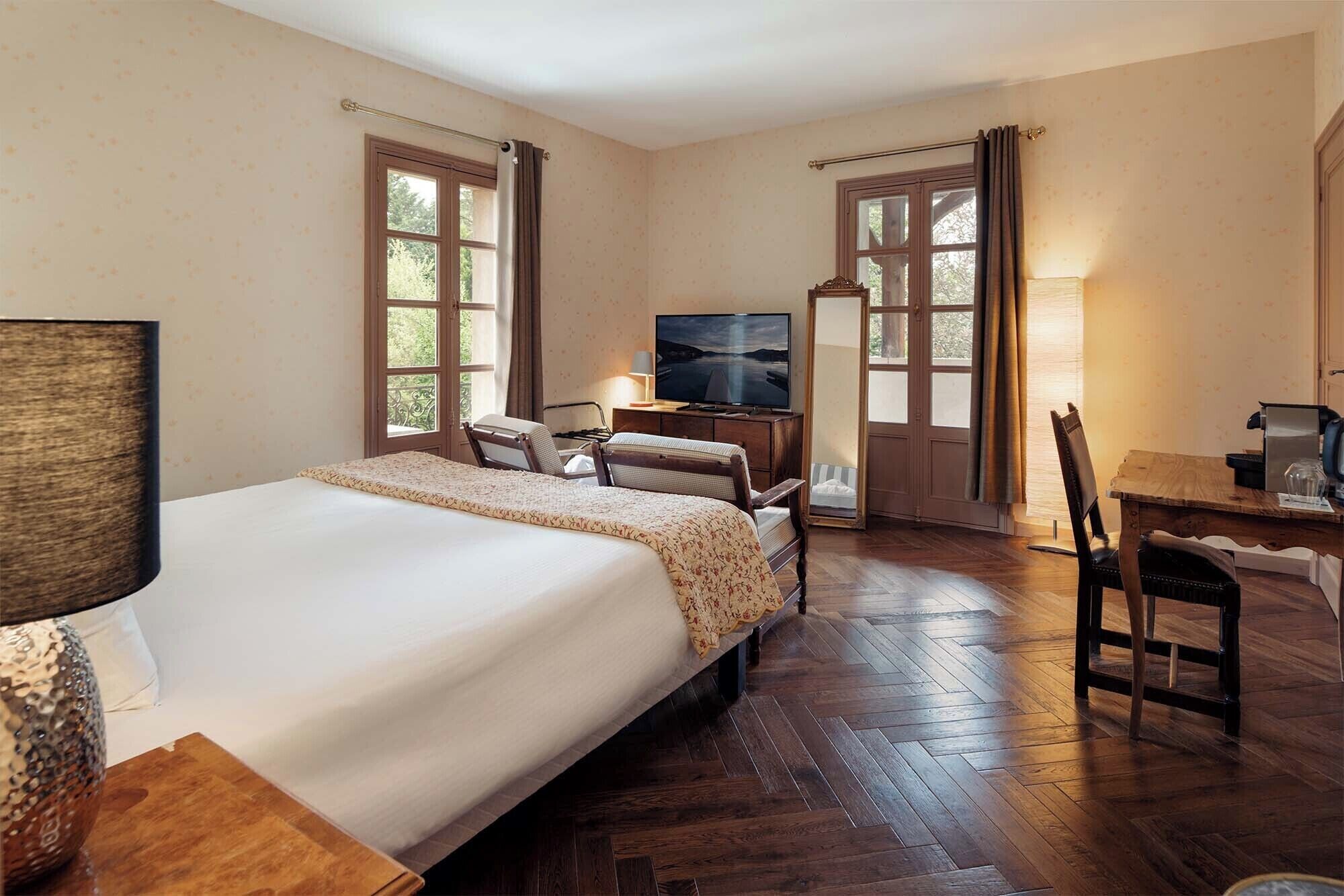 A hotel room with a large bed, a desk with a chair, a television, and two windows with curtains.