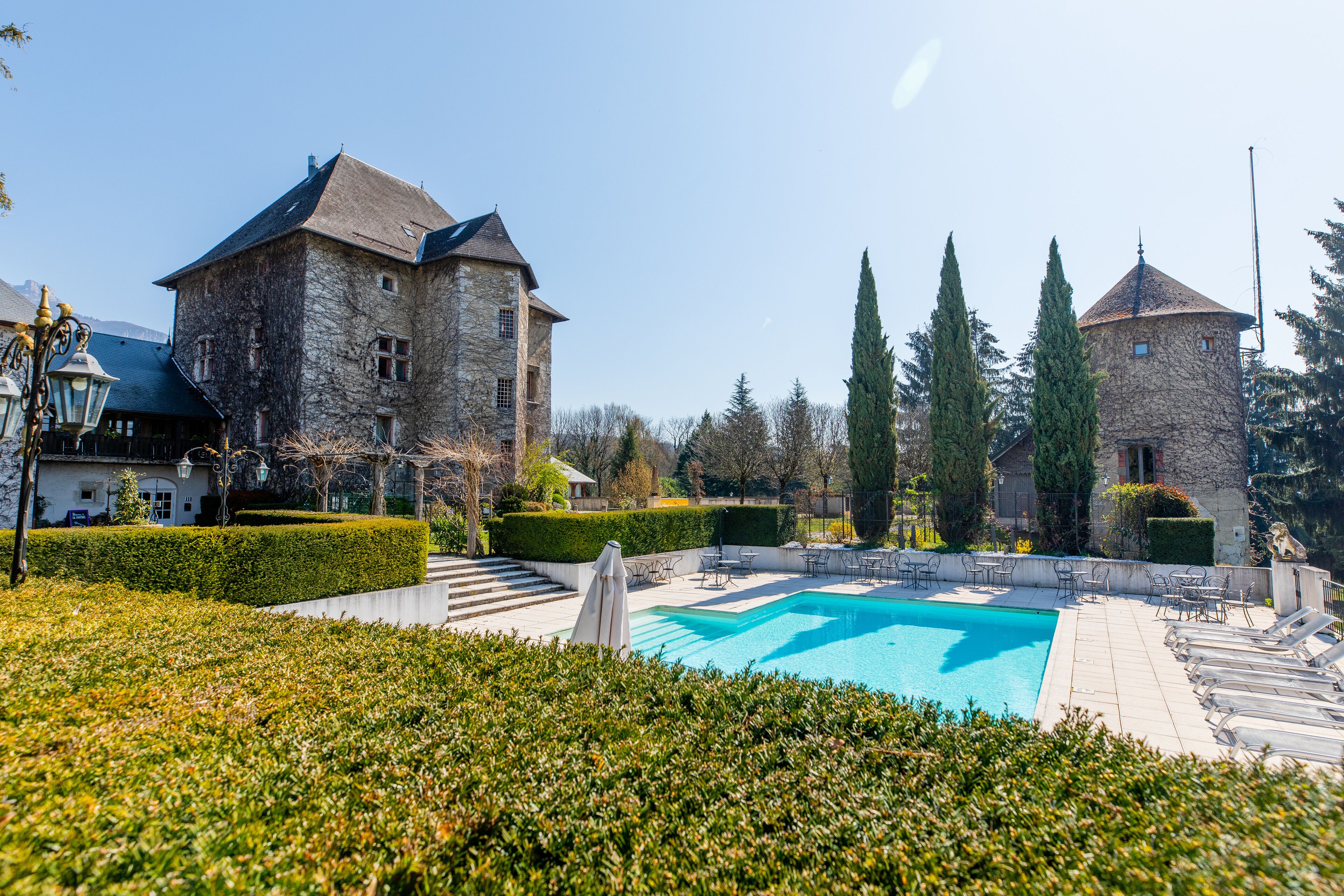 A stone building with a swimming pool, surrounded by hedges and trees.