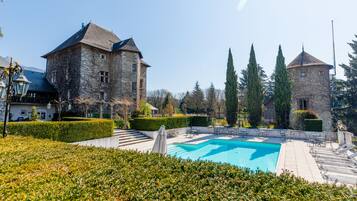 A stone building with a swimming pool, surrounded by hedges and trees.