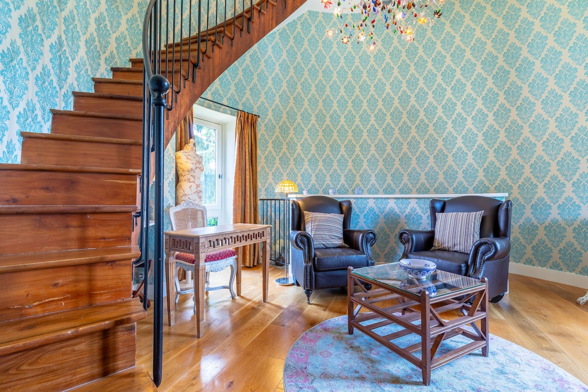 Una escalera con escalones de madera y barandilla negra, una sala de estar con papel tapiz estampado, dos sillones de cuero, una mesa de centro de vidrio y una mesa de comedor de madera con sillas.