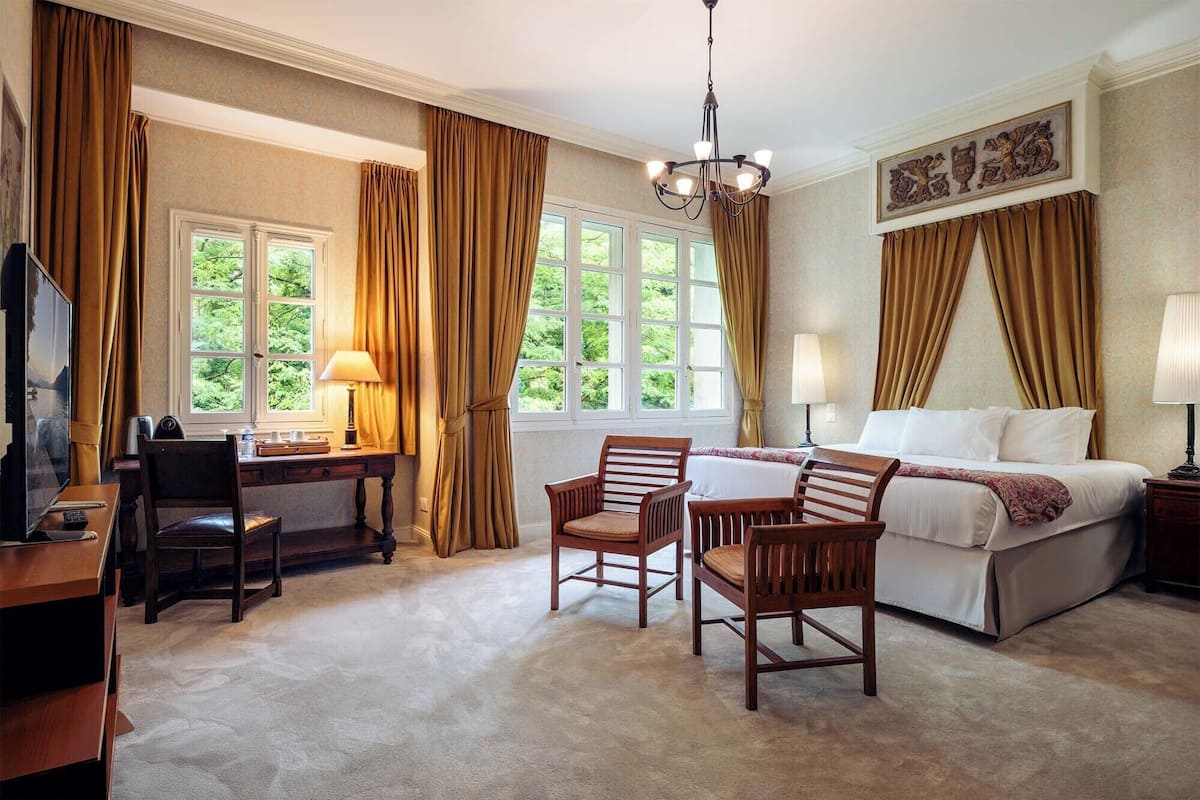 Habitación de hotel con cama, dos sillas, escritorio, televisor y ventanas grandes con cortinas.