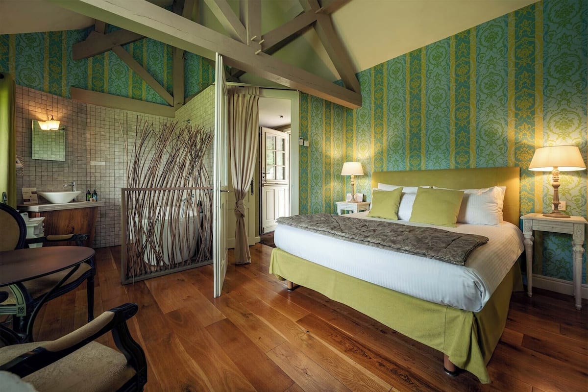 Un dormitorio con cama, mesitas de noche, una ventana y un baño con lavamanos y espejo.