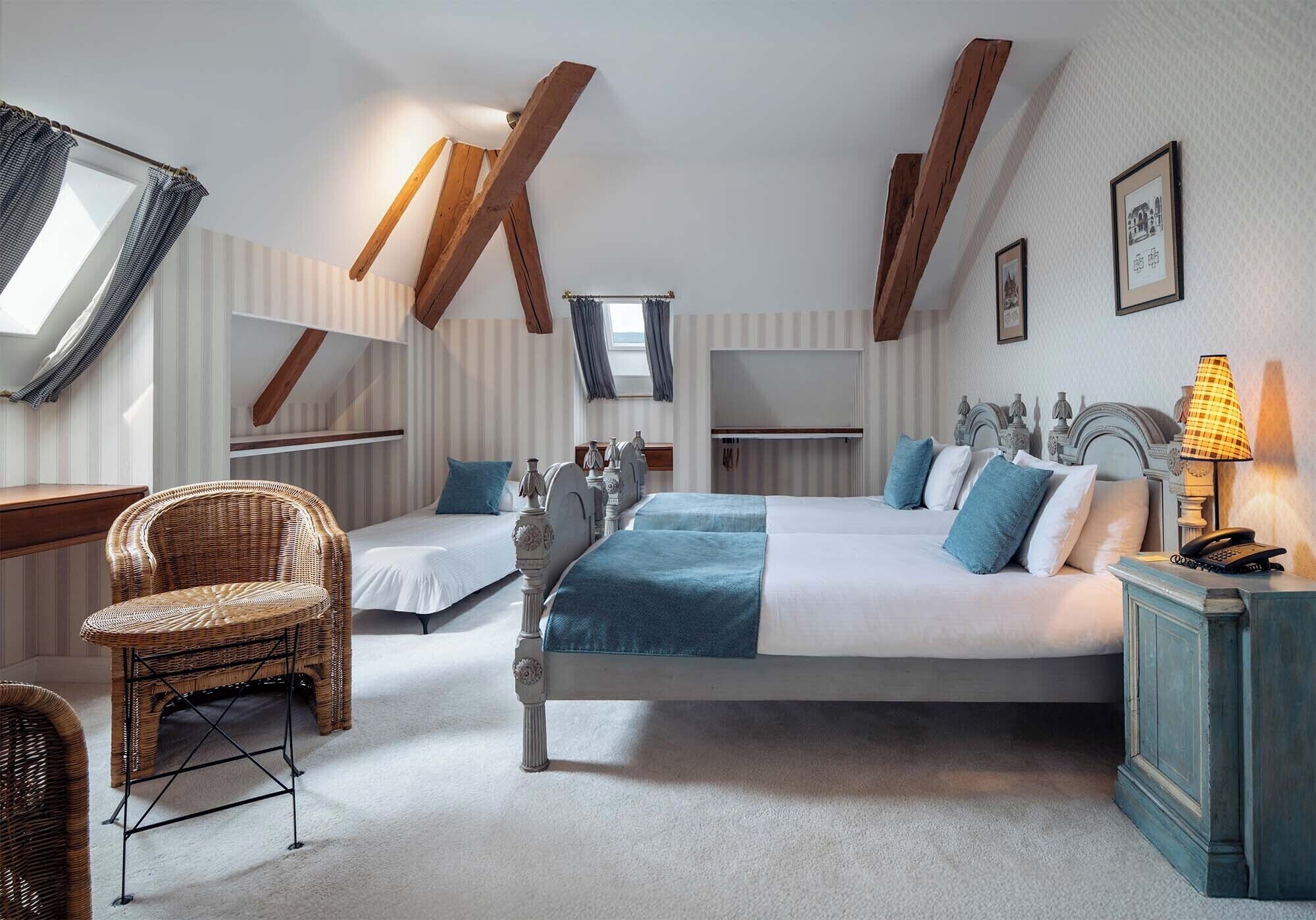 A bedroom with a large bed, two smaller beds, a wicker chair, a bedside table with a lamp, and a window with curtains.
