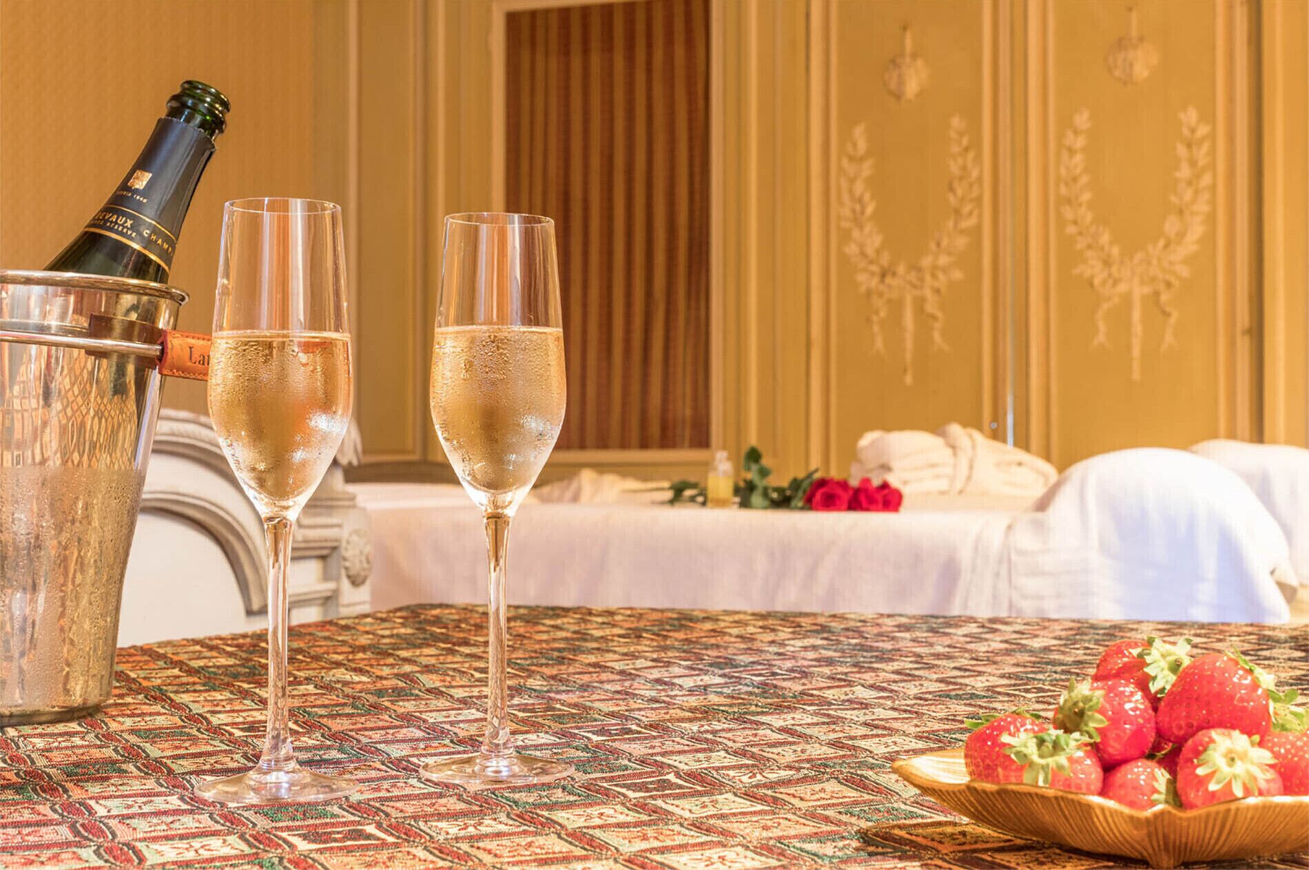Een kamer met een bed, een fles champagne, twee glazen en een schaal aardbeien op een tafelkleed met dessin.