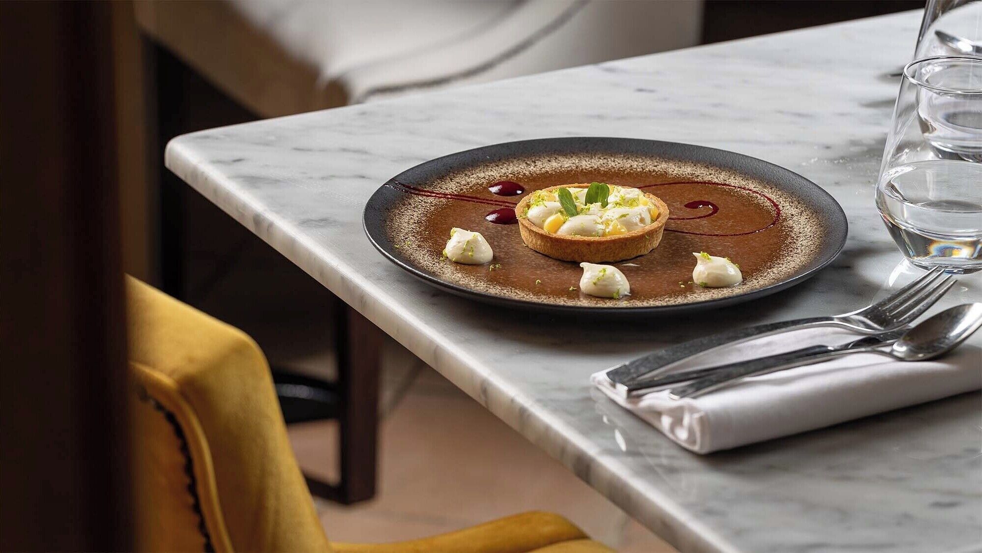 A dessert on a plate with a spoon and a glass of water on a marble table.
