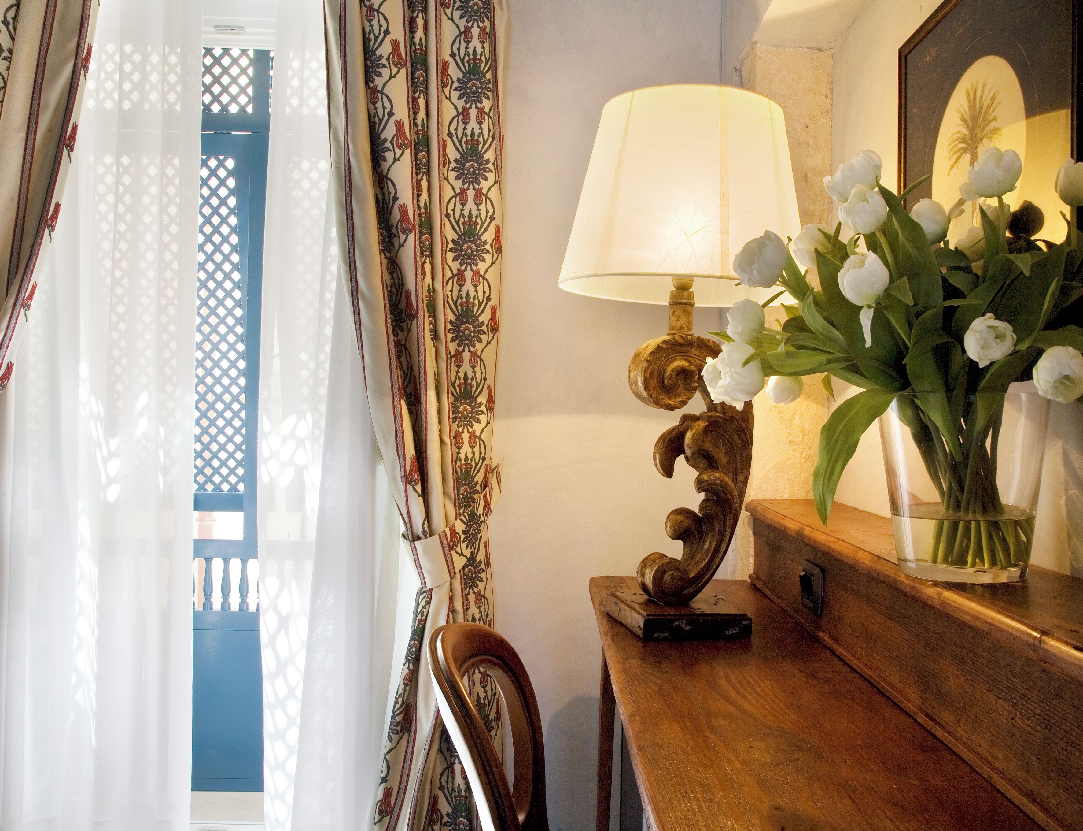 Ein Zimmer mit einem Holztisch, einer Lampe, einer Vase mit weißen Blumen und Blick durch ein Fenster mit gemusterten Vorhängen.