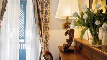 Ein Zimmer mit einem Holztisch, einer Lampe, einer Vase mit weißen Blumen und Blick durch ein Fenster mit gemusterten Vorhängen.