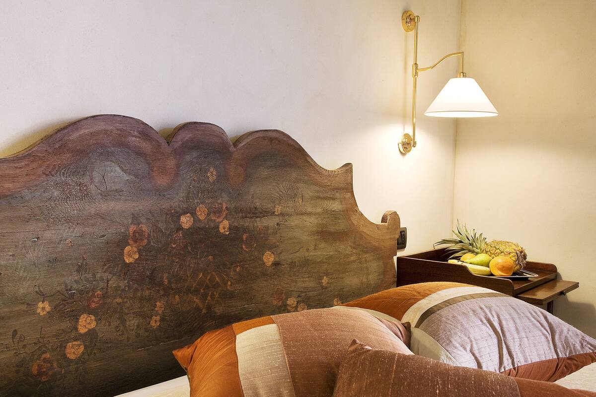 Un cabecero de madera con un diseño único, una mesita de noche con fruteros y una lámpara fijada a la pared.