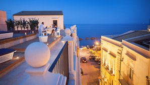 Una terraza en la azotea con mobiliario exterior, vista al mar y coches aparcados.