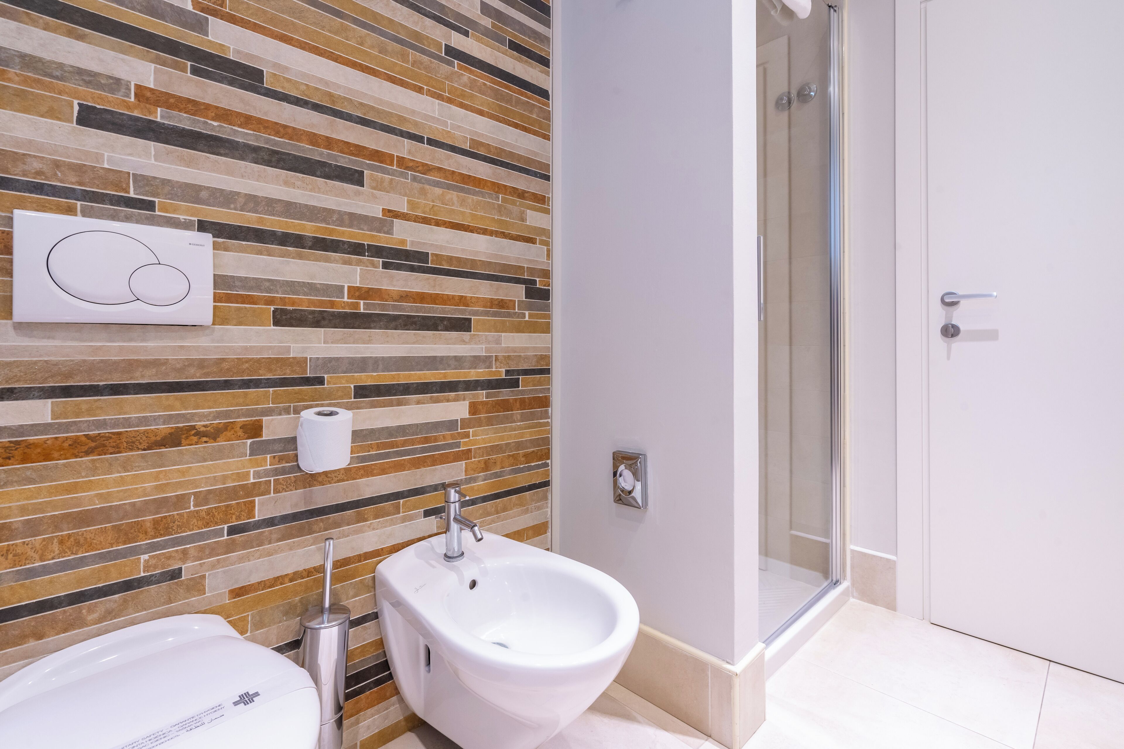 Une salle de bain dotée d’un revêtement mural moderne en carreaux, d’un bidet, d’un porte-rouleau de papier toilette et d’un espace douche.