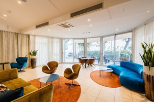 Eine moderne Hotelhalle mit einem gelben Sofa, einem blauen Sessel und einem orangefarbenen Teppich. Zwei runde Tische und ein blaues Sofa bieten Blick ins Freie.