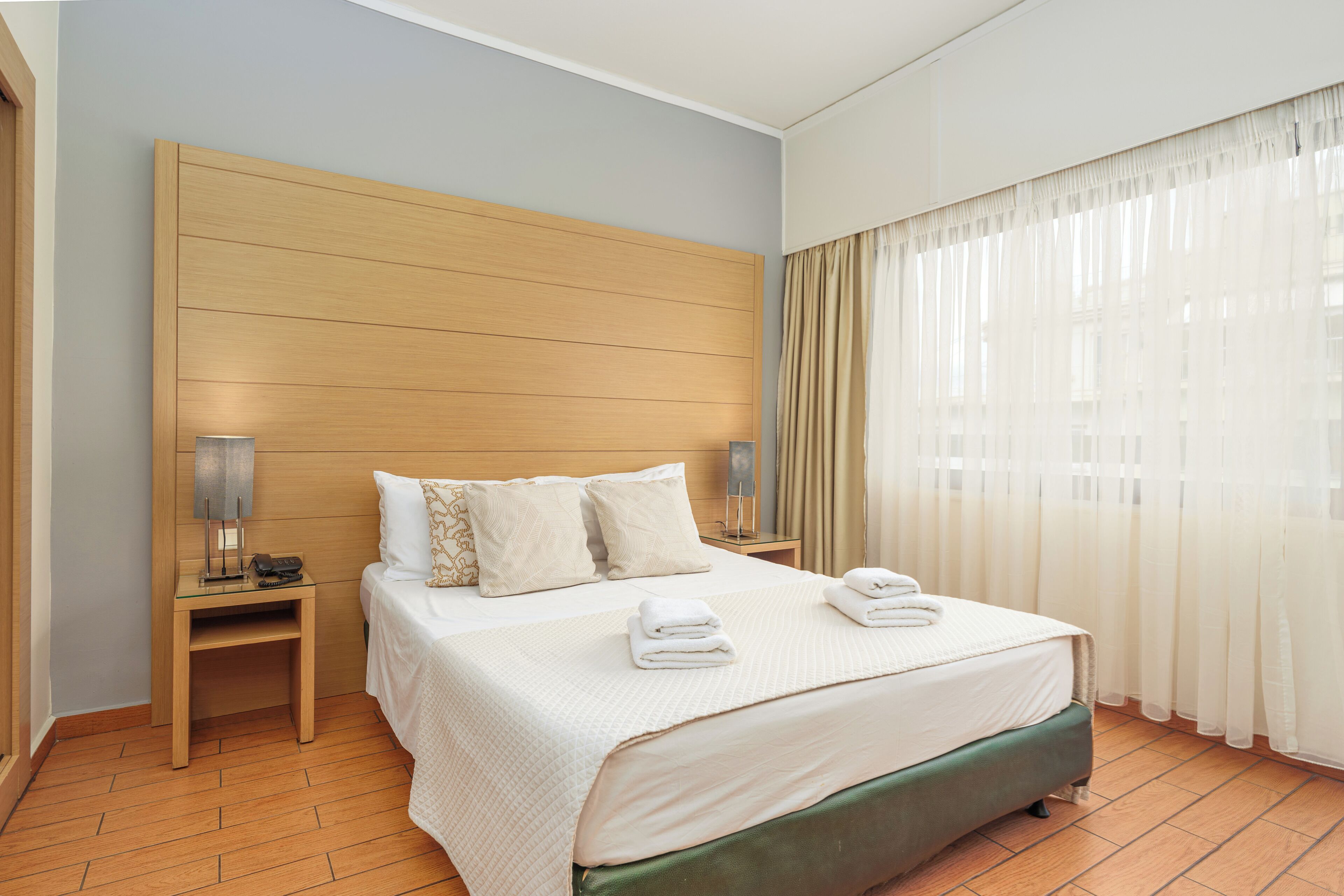 A neatly made bed with pillows, a bedside table with a lamp, and a window with sheer curtains.