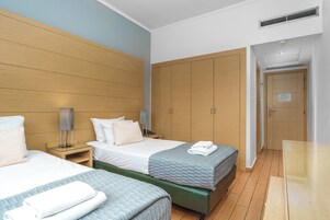 Quarto de hotel com duas camas, cabeceira de madeira, um guarda-roupa espaçoso e uma pequena mesa de cabeceira com abajur.