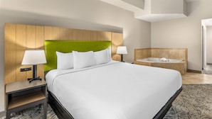 Une chambre d’hôtel avec un grand lit, deux lampes de chevet, une tête de lit verte, une table de chevet en bois et une salle de bain séparée avec une baignoire.