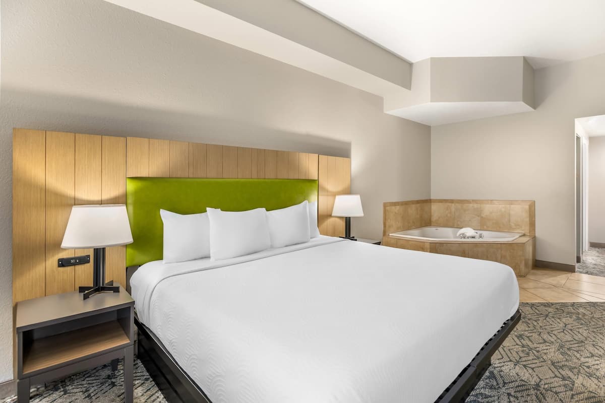 A hotel room with a large bed, two bedside lamps, a green headboard, a wooden nightstand, and a separate bathroom with a bathtub.