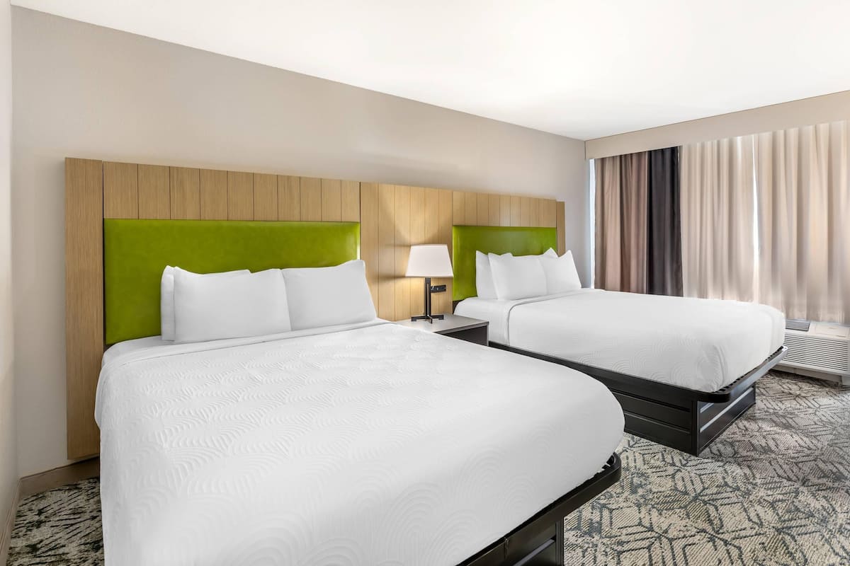 A hotel room with two beds, a wooden headboard, a nightstand with a lamp, and a large window with curtains.