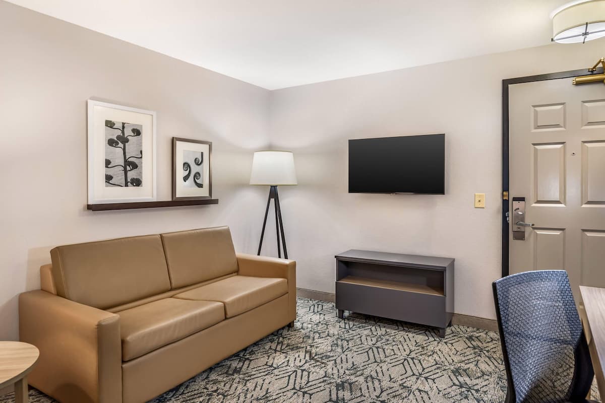 A hotel room with a beige sofa, a small table, a TV mounted on the wall, and a lamp.