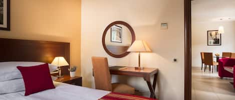 A hotel room with a bed, a desk, a chair, and a dining area visible through an open door.