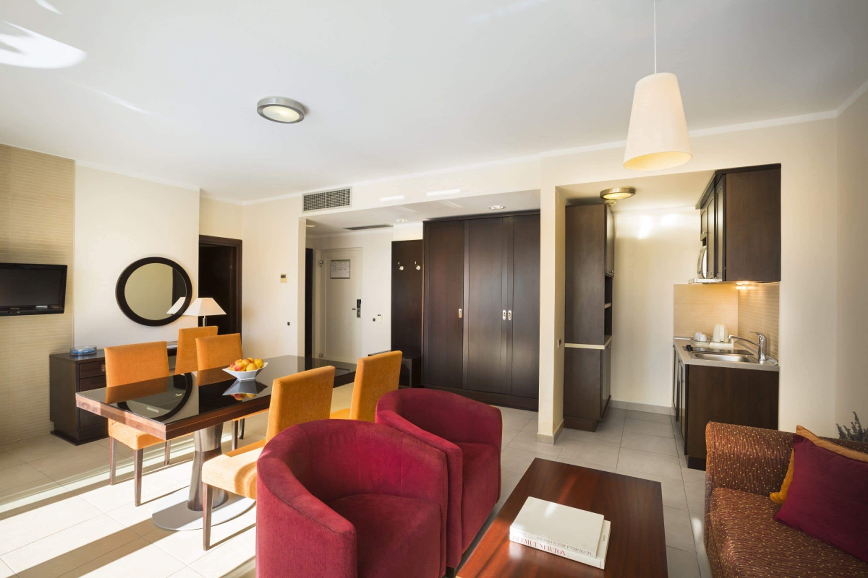 Une chambre d’hôtel moderne comprenant un coin repas, un salon avec des fauteuils rouges, un coin cuisine et un téléviseur fixé au mur.