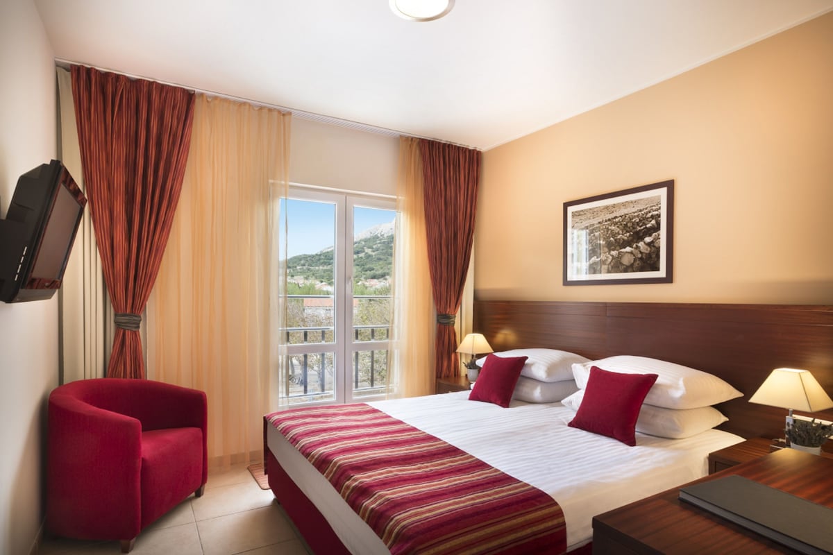 A hotel room with a large bed, a red armchair, a television, and a window with curtains.