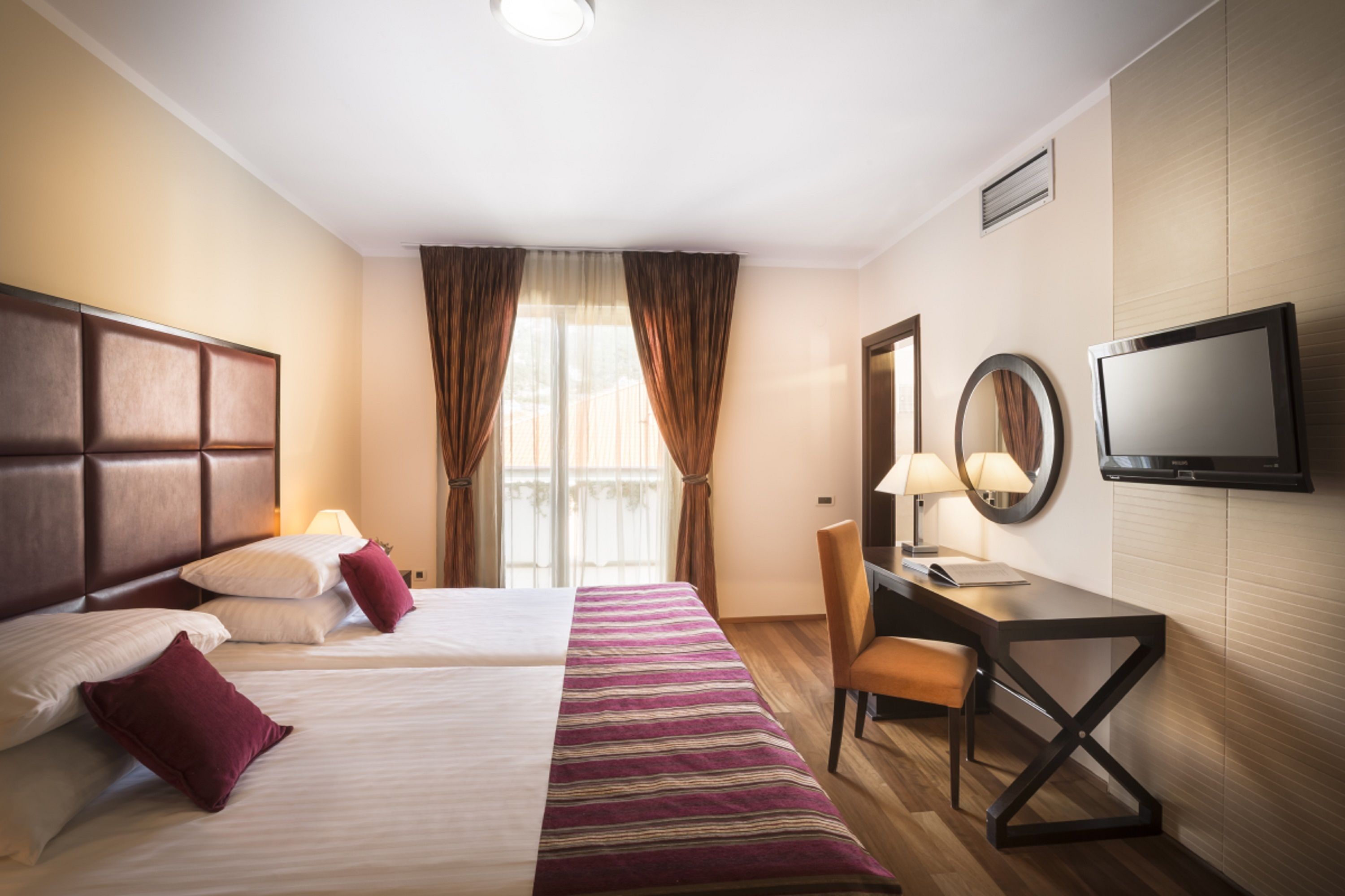 A hotel room with two beds, a desk, a chair, a TV, and a window with curtains.