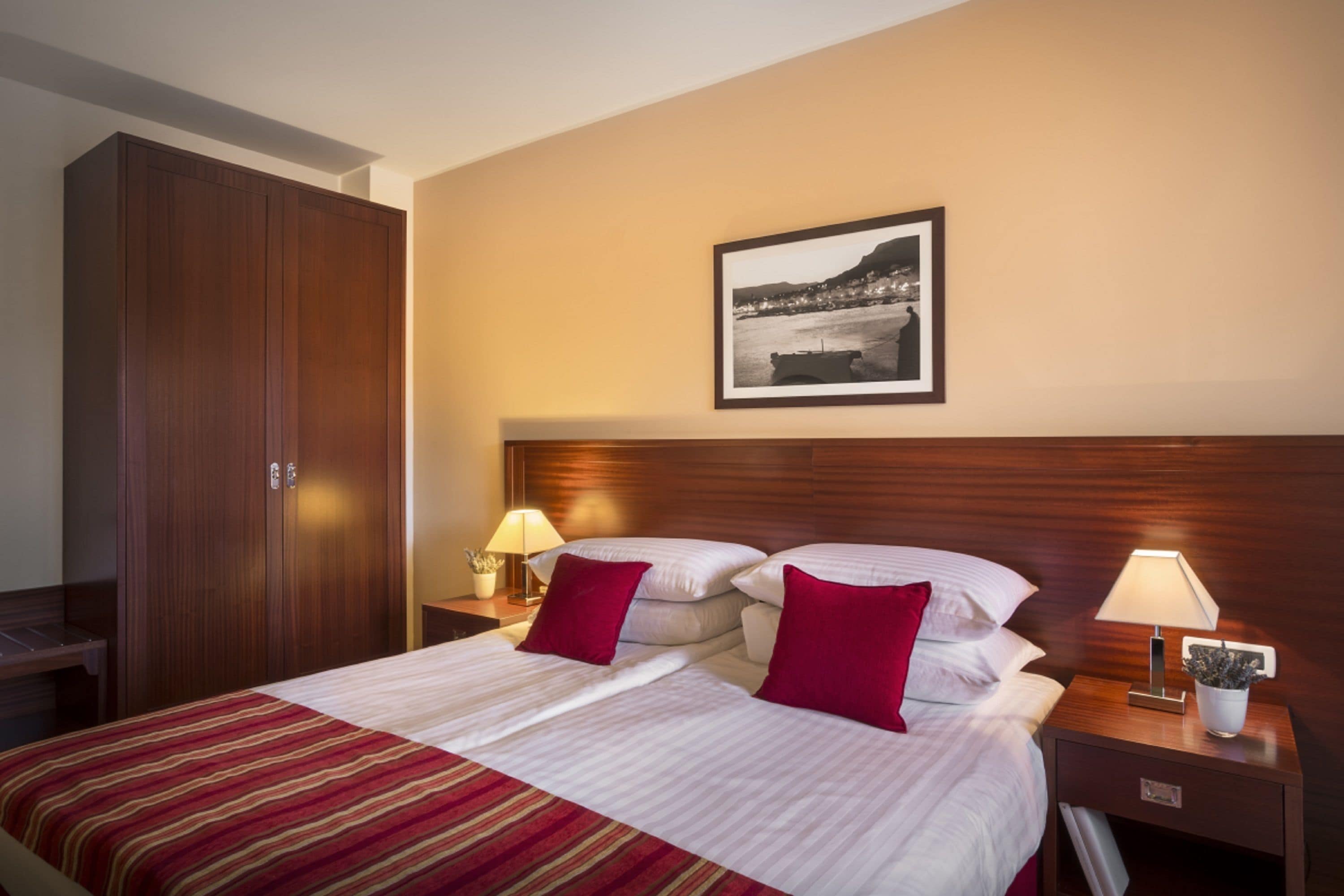 A hotel room with a large bed, two bedside tables with lamps, a wooden wardrobe, and a framed picture on the wall.