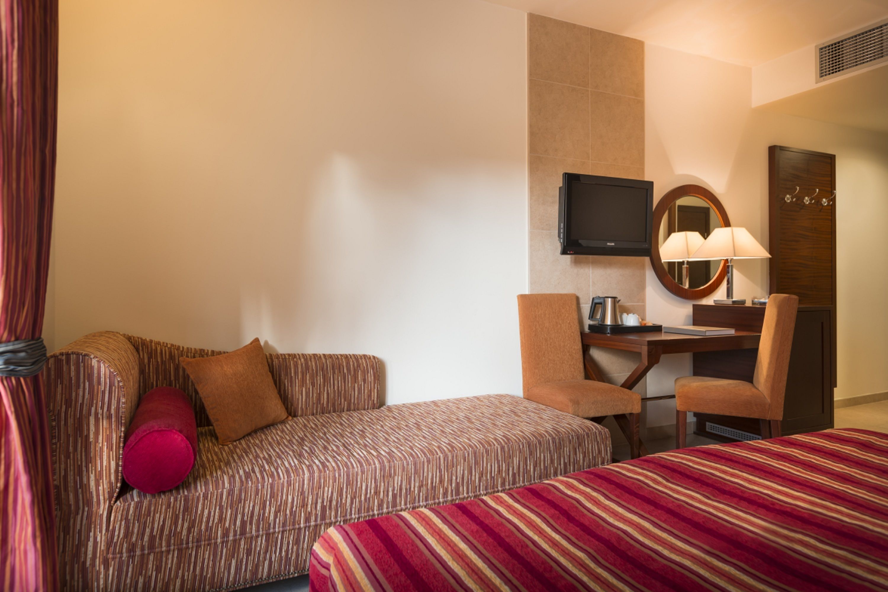 A hotel room with a sofa, a desk, a chair, a TV, a mirror, and a bed.