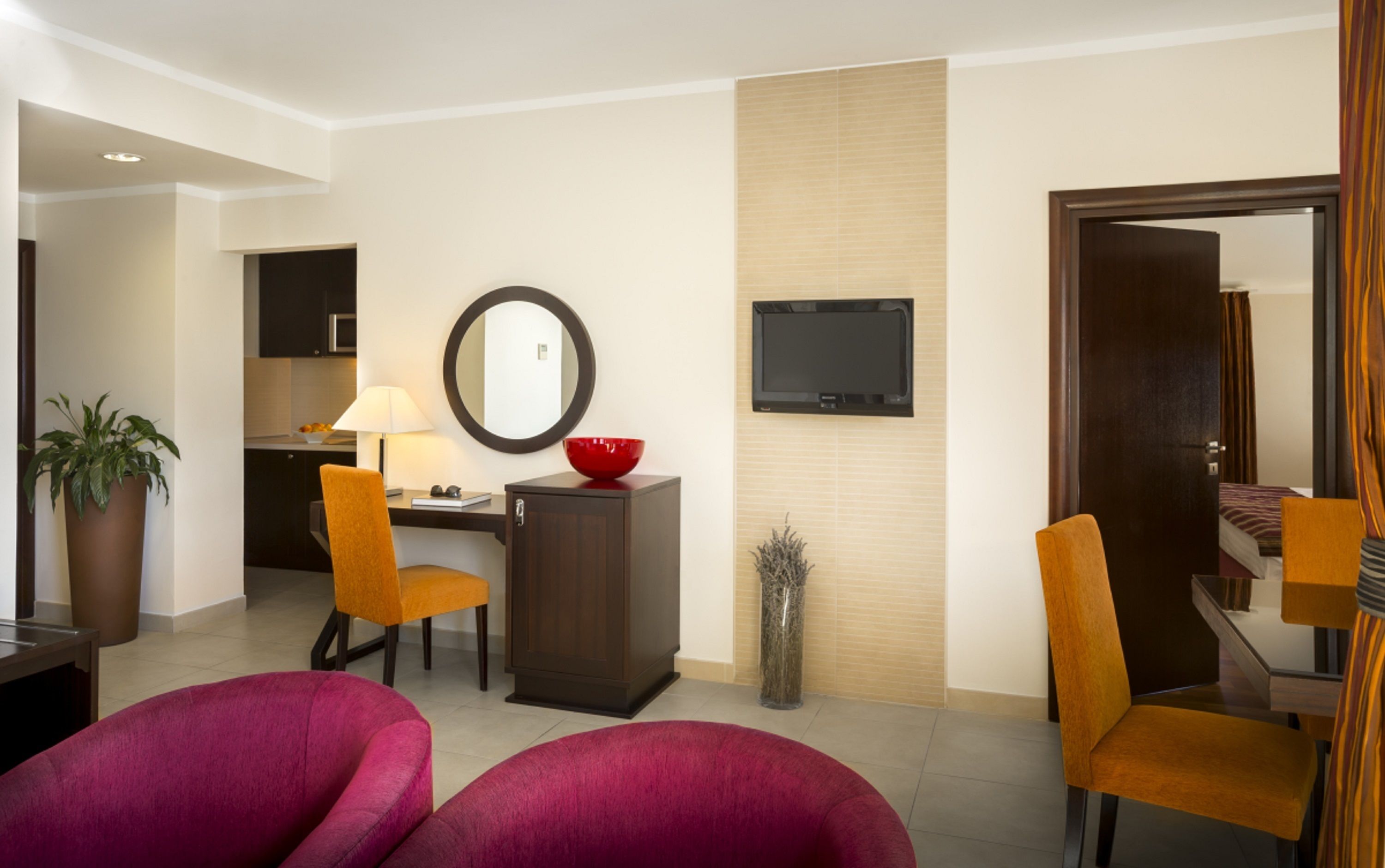A modern hotel room with a dining area, a desk, a chair, a red bowl, a mirror, and a TV.
