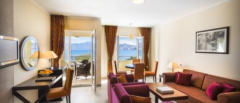 Quarto de hotel com sofá, cadeiras, mesa de jantar, televisão e vista para o mar.