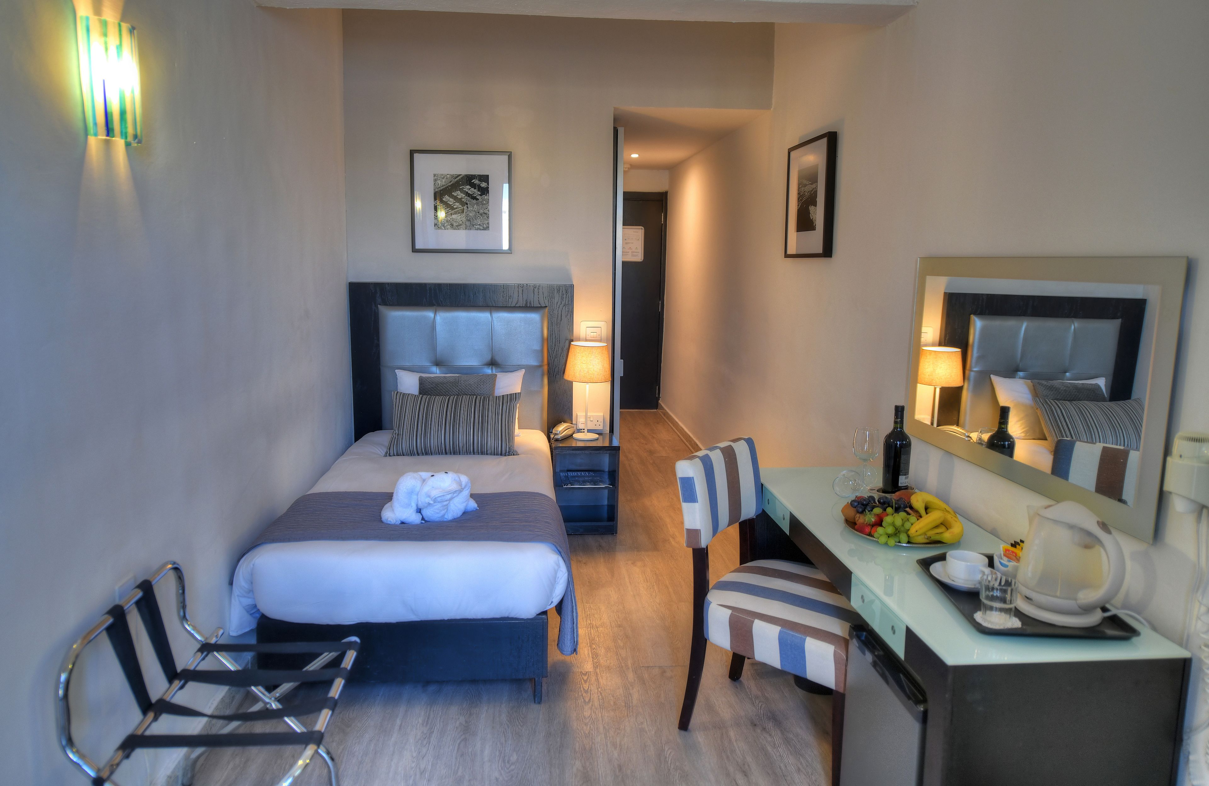 A hotel room with a double bed, a desk with a chair, a small table with a bowl of fruit, and a mirror above the desk.