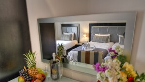 A hotel room with a large mirror reflecting the bed and headboard, a fruit bowl with various fruits, a bottle of wine, and a vase of flowers on the bedside table. - St Julian's Bay Hotel (St. Julian's)
