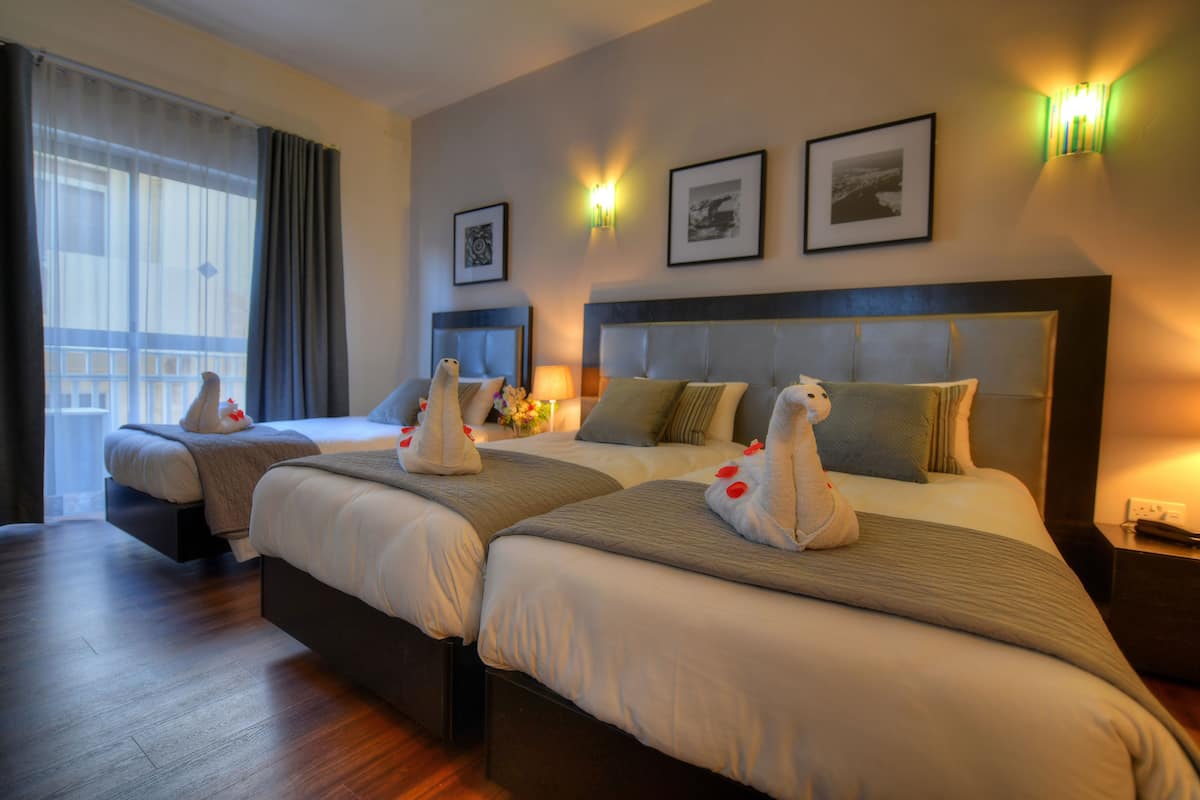 A hotel room with two beds, a large window with curtains, wall-mounted artwork, and a nightstand with a lamp.